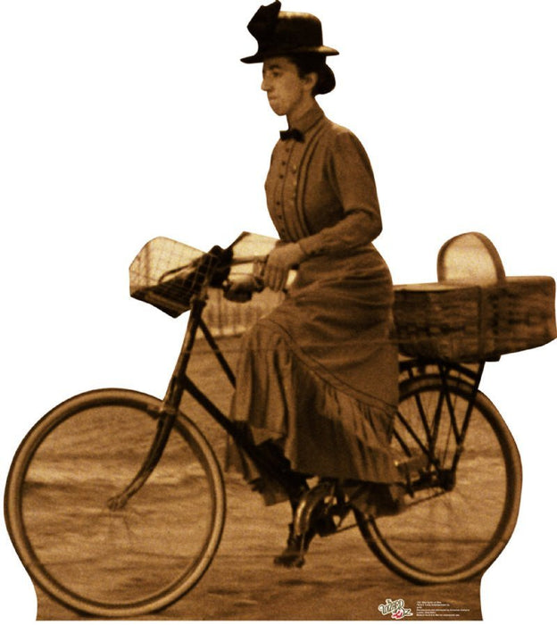 Life-size cardboard cutout in sepia tone of a woman in vintage attire riding a bicycle. She wears a long dress with buttons down the front, a wide-brimmed hat with a bow, and is carrying a large basket behind her on the bike.
