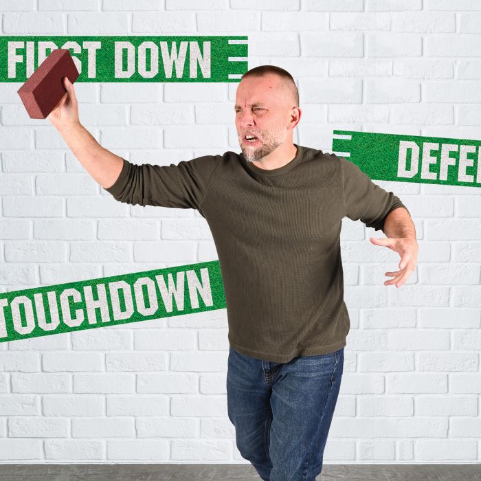 Man in a green sweater and jeans pretending to throw a foam brick, with a white brick wall backdrop decorated with green football-themed banners reading 'FIRST DOWN,' 'TOUCHDOWN,' and 'DEFENSE.'