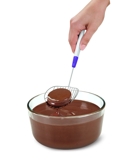  A hand holding a dipping tool with a round wire grid, lifting a chocolate-covered treat from a glass bowl filled with melted chocolate.
