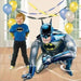 Decorative Batman-themed balloon featuring a young child wearing a blue Batman shirt, standing beside a large Batman balloon figure in a crouched pose, surrounded by blue, yellow, and white balloons and streamers in a bright room.