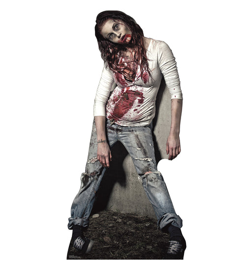 Life-size cardboard cutout of a female zombie with pale skin, blood-stained mouth and clothes, slumped against a concrete wall. She wears a torn white shirt and ripped jeans, with her head tilted unnaturally to the side.