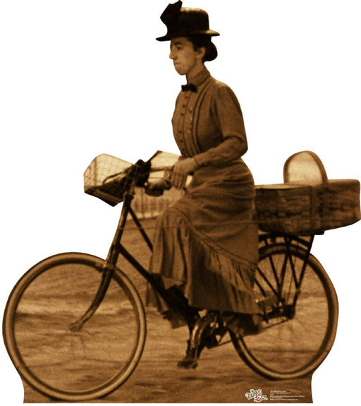 Life-size cardboard cutout in sepia tone of a woman in vintage attire riding a bicycle. She wears a long dress with buttons down the front, a wide-brimmed hat with a bow, and is carrying a large basket behind her on the bike.