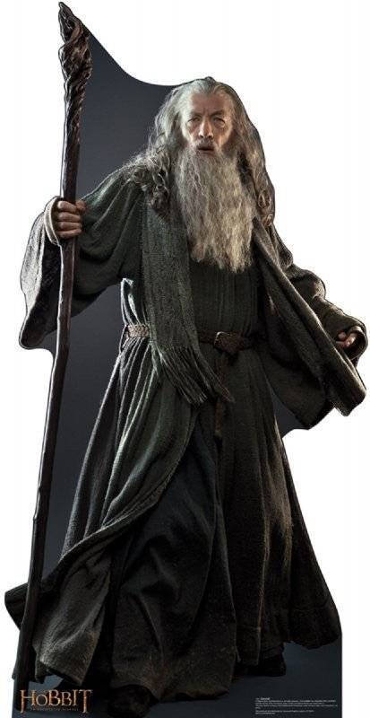 Life-size cardboard cutout of an elderly wizard with a long grey beard and flowing hair, dressed in a dark grey robe tied with a rope belt. He holds a tall wooden staff and stands in a powerful, commanding stance. The Hobbit movie logo is shown at the bottom corner.