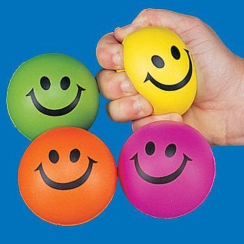 Four colorful smiley face stress balls in green, yellow, orange, and pink, with one being squeezed in a hand against a blue background.