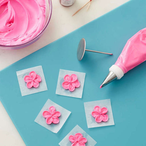 Piping setup on a blue surface showing a piping bag with pink icing and metal tip, a flower nail, and five parchment squares each with a piped pink icing flower. A bowl of pink icing and small brushes are also partially visible.
