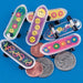 Set of six mini finger skateboards with white trim and black wheels, each featuring colorful designs like yellow smiley faces on purple, peace signs on green and blue, and multicolored flowers on pink, yellow, and lavender decks. U.S. coins shown for scale.
