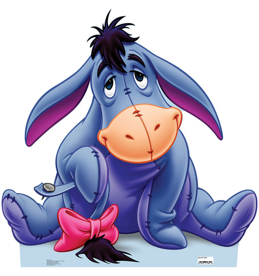 Cardboard cutout of Eeyore from Winnie the Pooh, seated with his legs splayed out and a melancholic expression. His body is a soft blue with visible stitched seams and his snout is a contrasting peach-orange. His long ears droop to either side, lined with a warm purple-pink. A black mane runs between his ears, and his detachable tail, pinned to his body, is accented by a bright pink bow. He rests on a soft blue shadow against a white background.