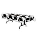 Rectangular table covered with a white tablecloth featuring a bold black cow print pattern, draped over a folding table.