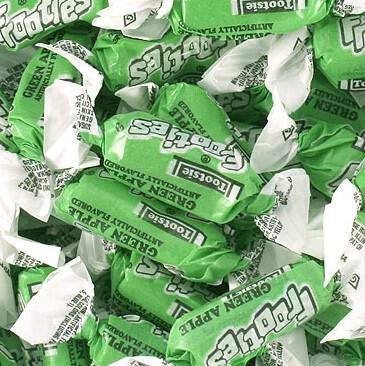 Close-up view of individually wrapped Tootsie Frooties Green Apple chewy candies with green and white wrappers, scattered in a pile.
