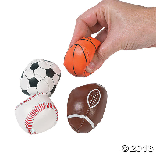Assorted foam sports balls, including a black and white soccer ball, an orange basketball with black lines, a brown football with white stripes and laces, and a white baseball with red stitching. A hand is shown squeezing the orange basketball.

