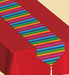 Colorful Latino table runner with vibrant horizontal stripes in red, orange, yellow, green, blue, and purple, placed over a solid red tablecloth, featuring a white tassel at the end.