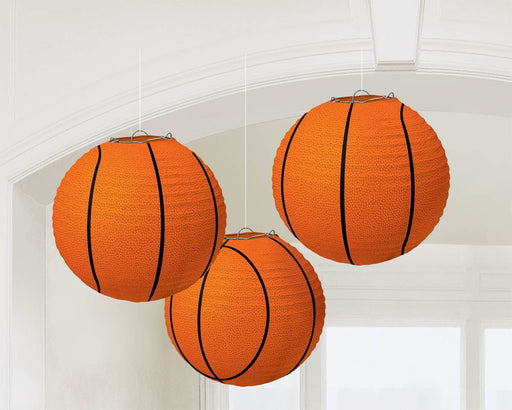 Three hanging paper lanterns designed to look like basketballs, with orange textured surfaces and black curved lines imitating the classic basketball pattern, suspended from the ceiling in a bright room.