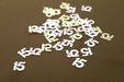 Scattered metallic confetti shaped like the number '15' on a brown surface, reflecting pale pink and white hues.