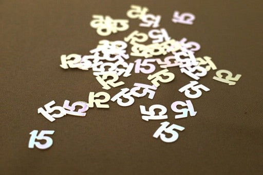 Scattered metallic confetti shaped like the number '15' on a brown surface, reflecting pale pink and white hues.