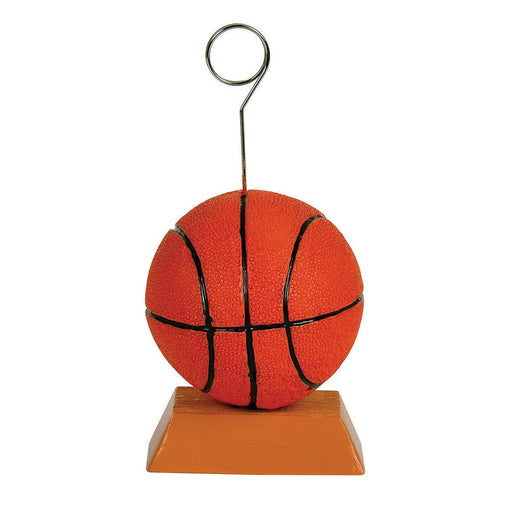Basketball-themed balloon or card holder featuring an orange basketball with black lines positioned on a brown rectangular base, with a vertical metal wire extending upward and ending in a circular clip for holding balloons, photos, or cards.