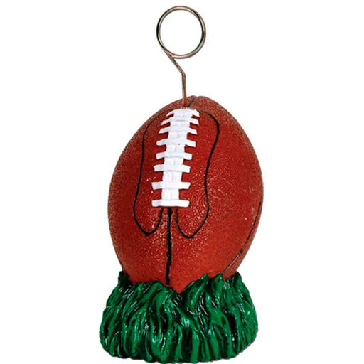 Football-shaped balloon weight featuring a brown football with white laces and black stitching, resting on a green grass-like base. A metal loop at the top is designed for securing balloons, perfect for sports-themed parties and events.