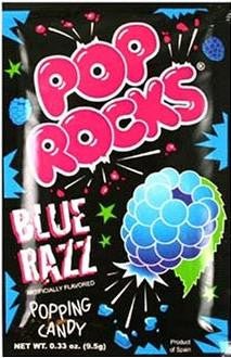A small black packet of Pop Rocks Blue Razz popping candy, featuring vibrant graphics of exploding stars and a blue raspberry illustration. The brand name 'Pop Rocks' is displayed in bold, pink, bubble-style letters, with 'Blue Razz' and 'Popping Candy' written below in white and blue fonts. Net weight is 0.33 oz (9.5g).
