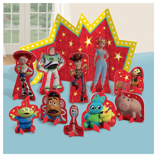 Toy Story-themed table centerpiece set featuring stand-up cutouts of characters including Woody, Buzz Lightyear, Jessie, Bo Peep, Hamm, Mr. Potato Head, Forky, the Aliens, Ducky, and Bunny. The backdrop is a large red starburst design with yellow lights and stars.